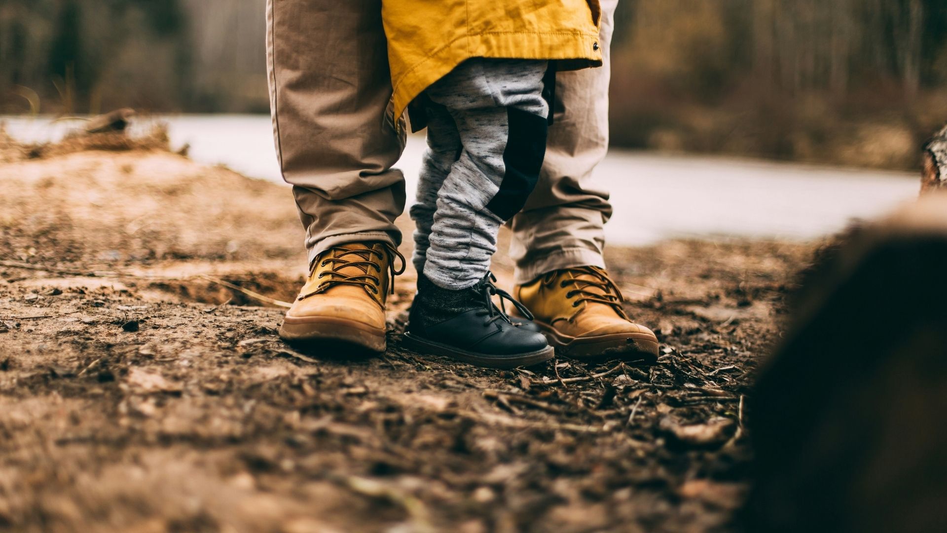 Das Bild zeigt die Füße eines Erwachsenen und eines Kindes, die nebeneinander stehen. Der Erwachsene trägt beige Hosen und braune Schnürstiefel, das Kind graue Hosen und schwarze Schnürschuhe.  Der Hintergrund ist unscharf und zeigt eine natürliche Umgebung, möglicherweise eine Flusslandschaft. Der Fokus liegt auf den Schuhen und vermittelt ein Gefühl von Nähe und Verbundenheit.